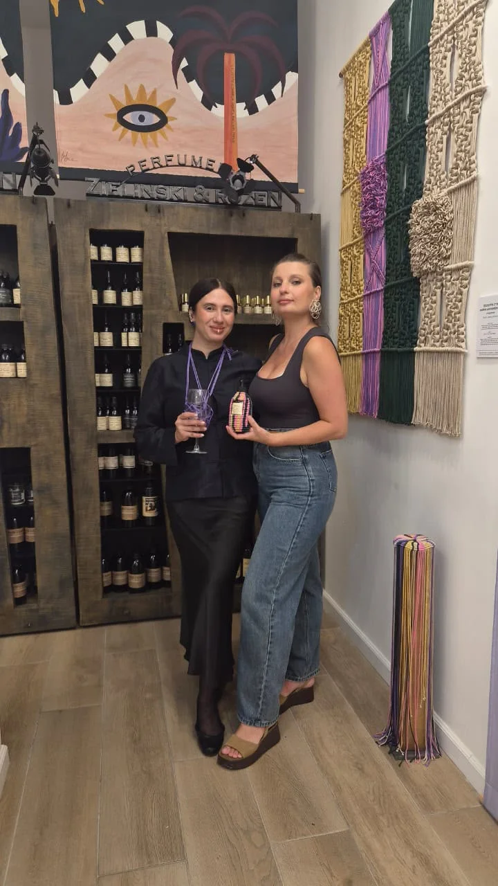 שתי משתתפות מחזיקות בקבוקי בושם מול קיר המקרמה ומדפי הבשמים