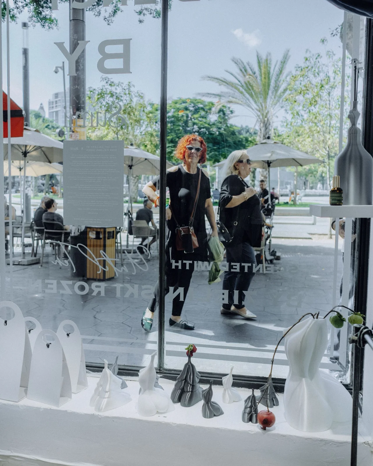 מבט מתוך הגלריה אל כיכר דיזינגוף — מבקרות חולפות מול חלון הראווה ופסלוני SUMII על מדף החלון